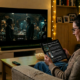 A person wearing glasses and a beige jumper sits on a sofa, looking at a tablet in their hands while a large television screen displays a sci-fi movie scene with two characters in a dimly lit setting. The warm, cosy living room is illuminated by ambient lighting, with a bowl of popcorn on the coffee table.
