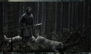 Aaron Taylor-Johnson, portraying a bearded medieval farmer in a dark period clothing, stands on a mossy log holding a long spear in a dense, desaturated forest alongside shaggy grey hunting dogs in Werwulf.