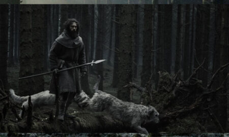 Aaron Taylor-Johnson, portraying a bearded medieval farmer in a dark period clothing, stands on a mossy log holding a long spear in a dense, desaturated forest alongside shaggy grey hunting dogs in Werwulf.