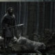 Aaron Taylor-Johnson, portraying a bearded medieval farmer in a dark period clothing, stands on a mossy log holding a long spear in a dense, desaturated forest alongside shaggy grey hunting dogs in Werwulf.