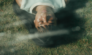 Isabel in Filipinana with long dark hair lying on green grass, seen from a high angle through a glass reflection, with their eyes closed and a calm expression, wearing a light blue and white trim sleeveless top.
