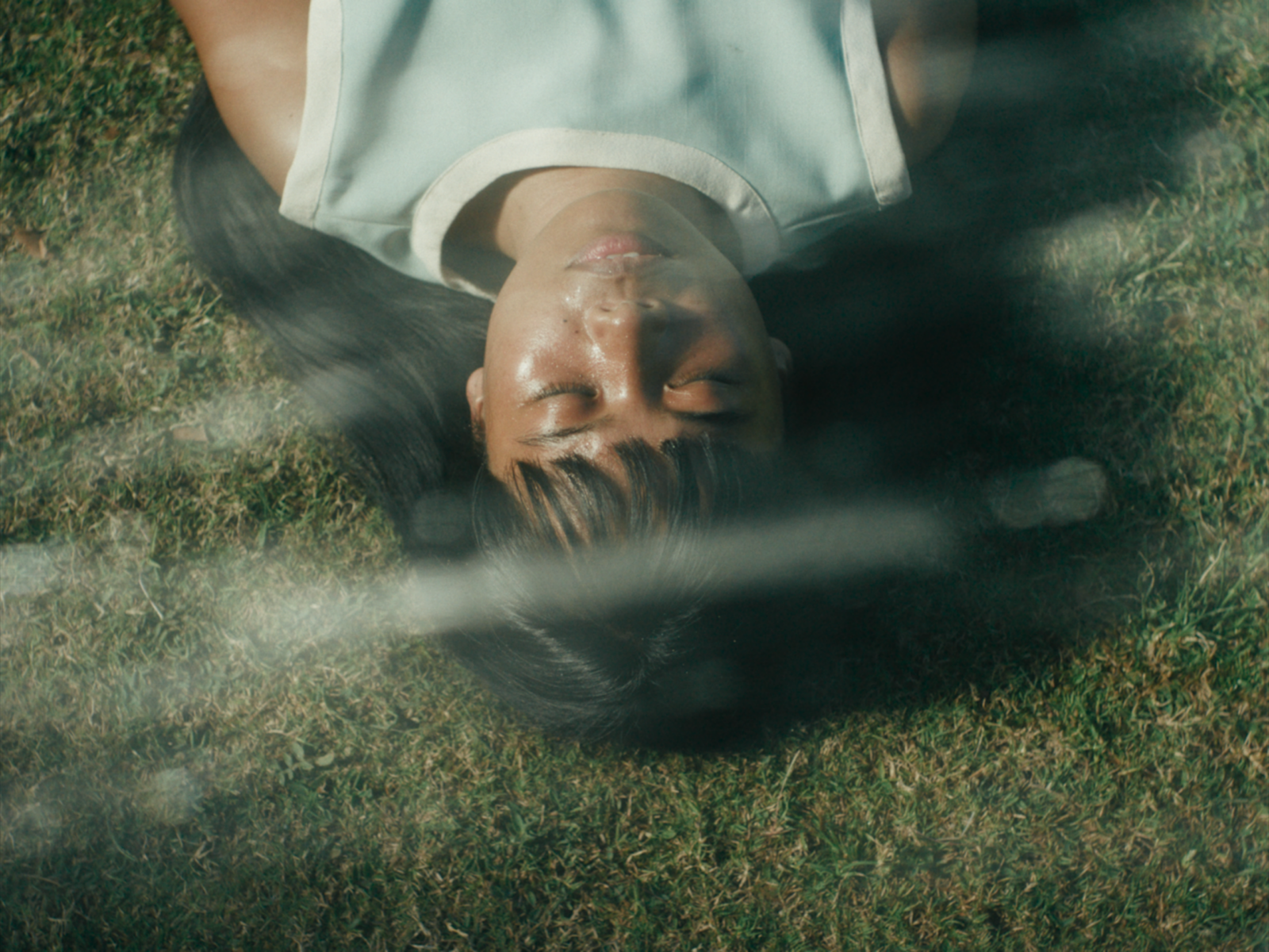 Isabel in Filipinana with long dark hair lying on green grass, seen from a high angle through a glass reflection, with their eyes closed and a calm expression, wearing a light blue and white trim sleeveless top.