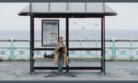 A person wearing a tan trench coat sits on a wooden bench inside a bus shelter by the sea, featuring a large dark fish-like tail instead of legs, while writing on a clipboard. The shelter displays a bus timetable panel reading "coaster BUS INFORMATION" - from the mermaid comedy A Tail.