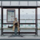 A person wearing a tan trench coat sits on a wooden bench inside a bus shelter by the sea, featuring a large dark fish-like tail instead of legs, while writing on a clipboard. The shelter displays a bus timetable panel reading "coaster BUS INFORMATION" - from the mermaid comedy A Tail.