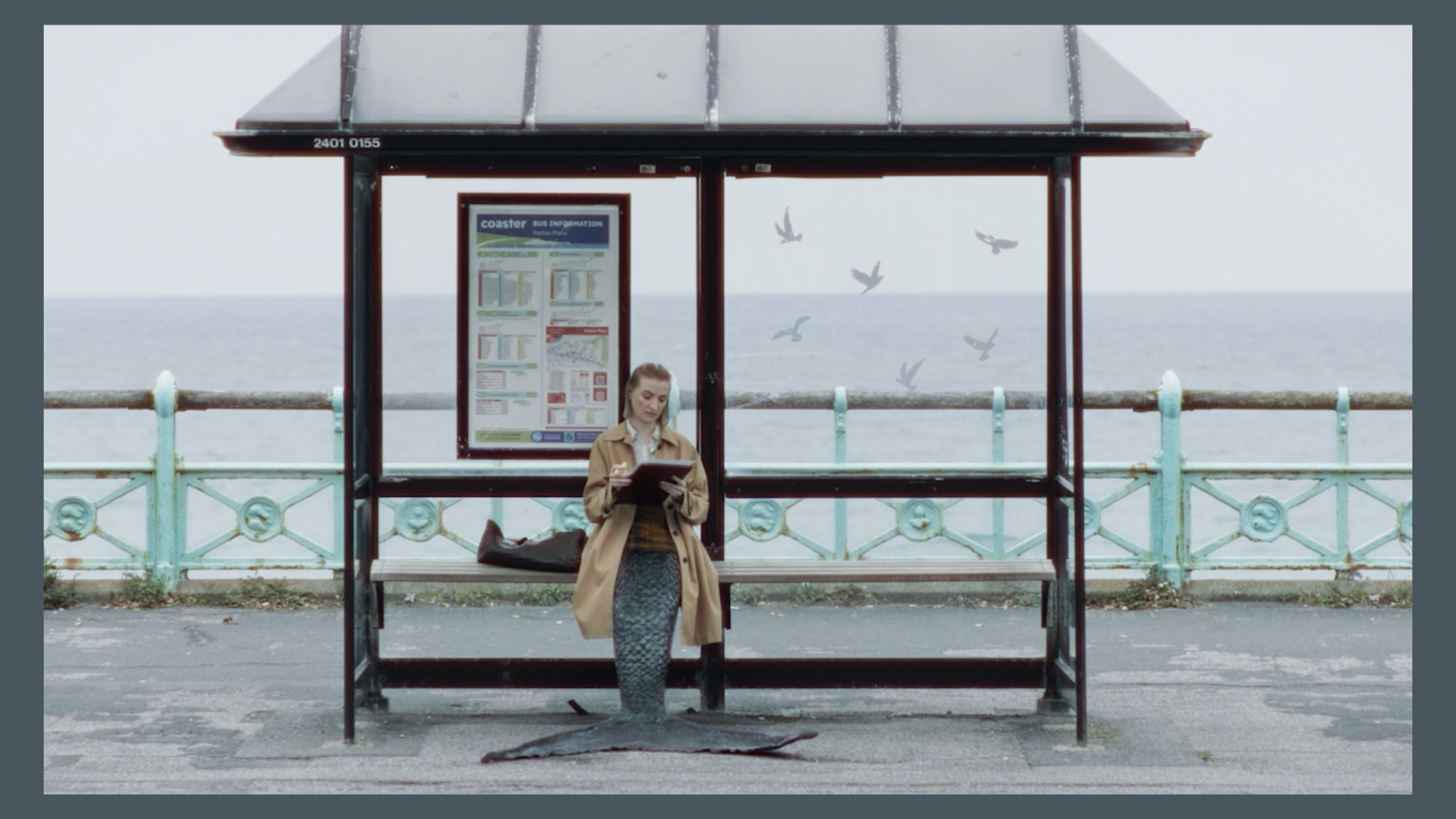 A person wearing a tan trench coat sits on a wooden bench inside a bus shelter by the sea, featuring a large dark fish-like tail instead of legs, while writing on a clipboard. The shelter displays a bus timetable panel reading "coaster BUS INFORMATION" - from the mermaid comedy A Tail.