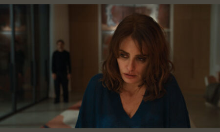 Penélope Cruz, playing Sofia in the film Bunker, looks downward with a troubled expression in a blue v-neck top, while a blurry Javier Bardem, as Miguel, stands in the background.