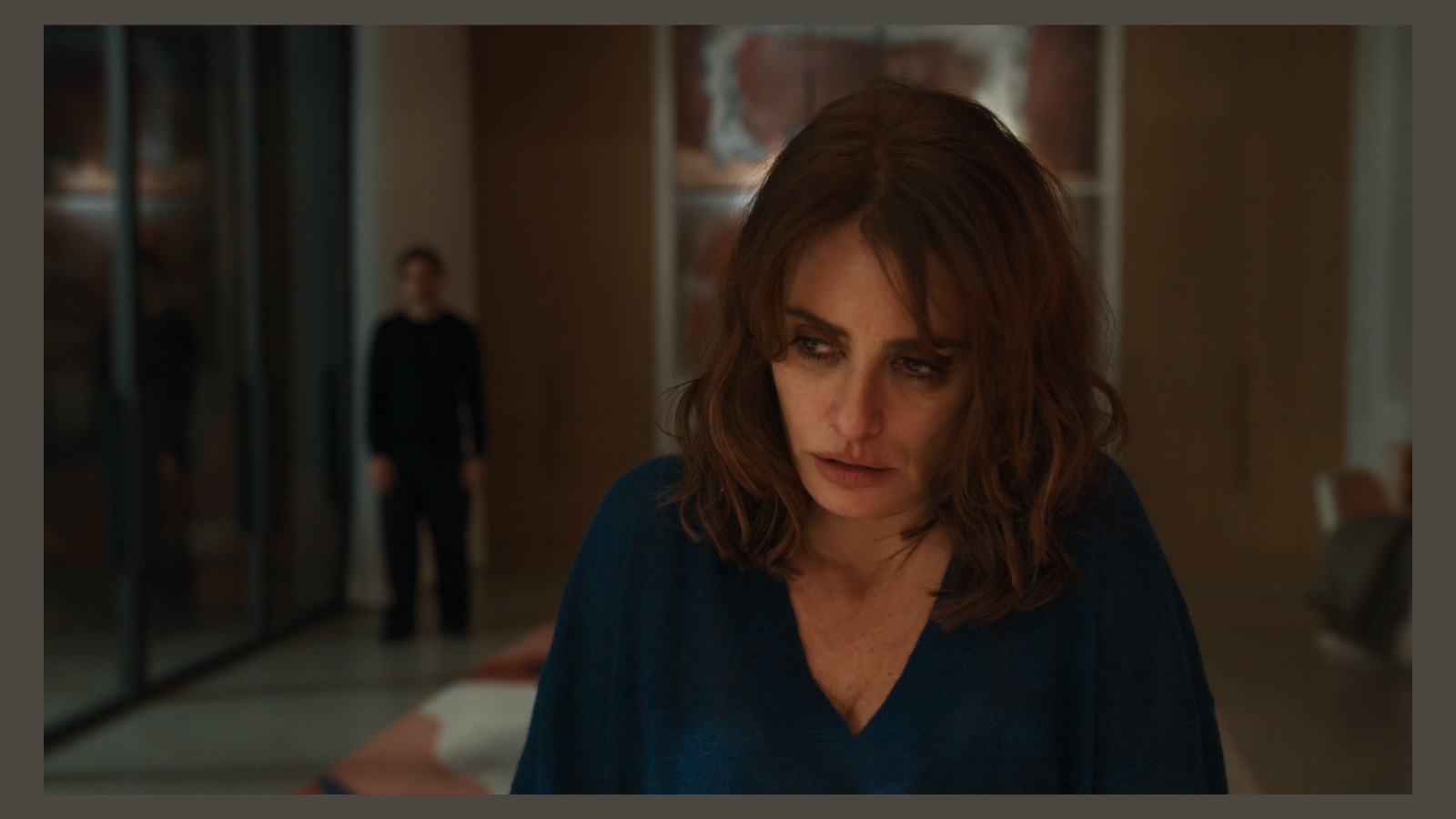 Penélope Cruz, playing Sofia in the film Bunker, looks downward with a troubled expression in a blue v-neck top, while a blurry Javier Bardem, as Miguel, stands in the background.